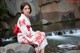 A woman in a red and white kimono sitting on a rock by a waterfall.